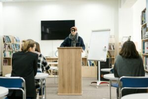 A man wearing sunglasses, a hat, and a scarf stands behind a podium in a library room with people sitting at desks in front of him.