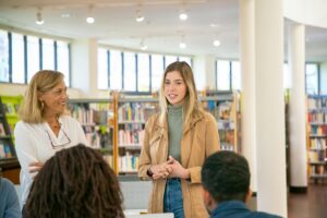 A young woman speaks in a library filled with shelves of books and windows in front of a group of people standing next to a smiling older woman.